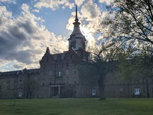 Historic building with a clock tower, surrounded by trees and a cloudy sky, illuminated by sunlight.