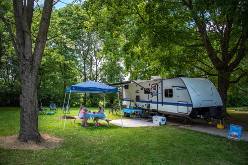 A camper trailer parked under trees, with two people sitting at a table and a blue canopy nearby.