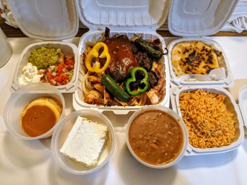 A takeout meal featuring grilled meats, rice, beans, guacamole, pico de gallo, and dessert in divided containers.