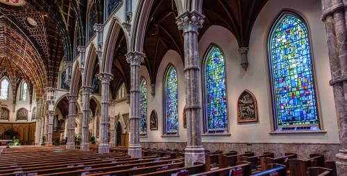 Interior of a church featuring tall stained glass windows and wooden pews, with ornate arches and a vaulted ceiling.
