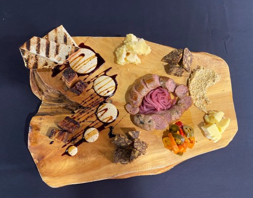 A wooden platter featuring assorted meats, cheeses, pickled vegetables, and grilled bread, drizzled with sauce.