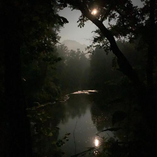 A serene night scene of a river reflecting the moonlight, framed by dark trees and misty mountains in the background.