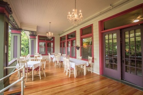 A bright dining area with white tables and chairs, chandeliers, and large windows overlooking greenery.