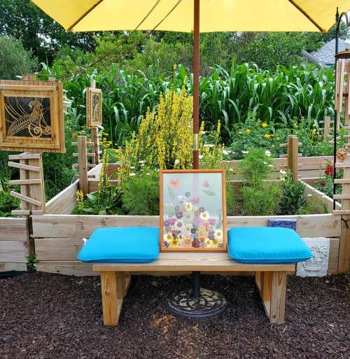 A wooden bench with blue cushions under a yellow umbrella, surrounded by a garden and a framed floral artwork.