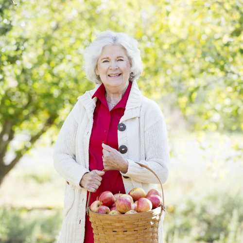 Smiling elderly woman in a cozy sweater holding a basket of apples, standing outdoors with greenery in the background.