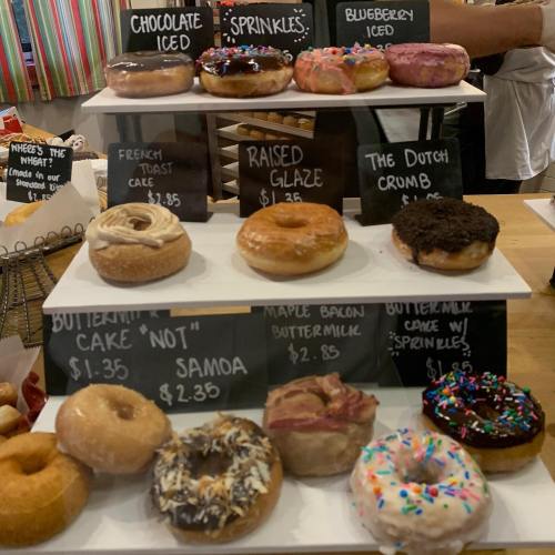 A display of assorted donuts with labels, including chocolate iced, blueberry iced, and maple bacon buttermilk.