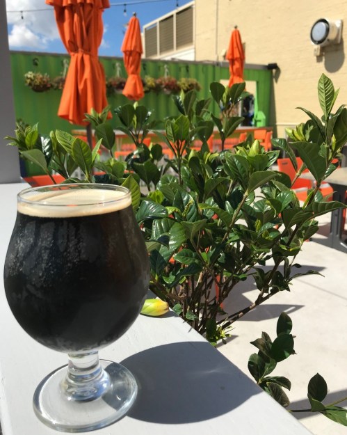 A dark beer in a glass sits on a table, with green plants and orange umbrellas in the background on a sunny day.