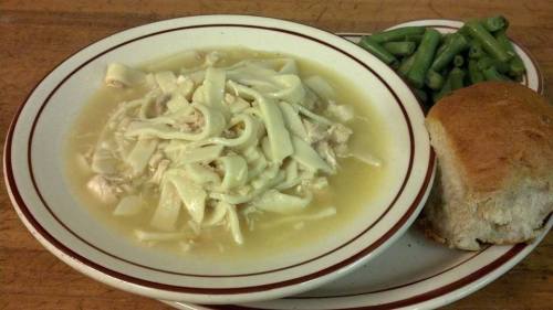 A bowl of chicken noodle soup with green beans and a piece of bread on a plate.