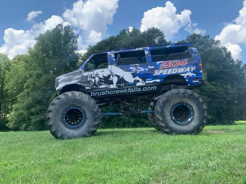 A large monster truck with a blue and gray design parked on green grass, surrounded by trees and a blue sky.