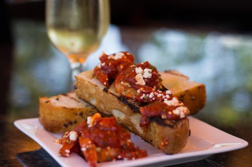 A plate with toasted bread topped with tomato sauce and feta cheese, alongside a glass of white wine.