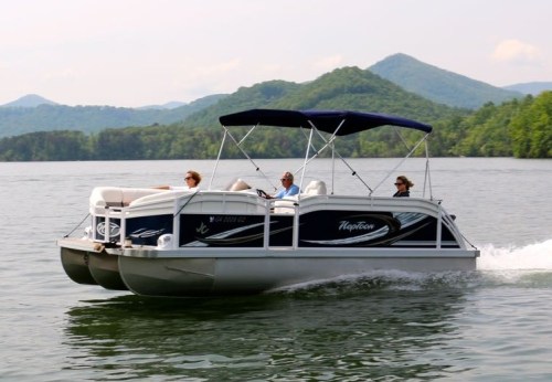 A pontoon boat with three people cruising on a lake surrounded by green mountains under a clear sky.