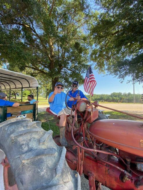 Two people on a vintage tractor, one holding an American flag, smiling and giving a thumbs up in a sunny outdoor setting.