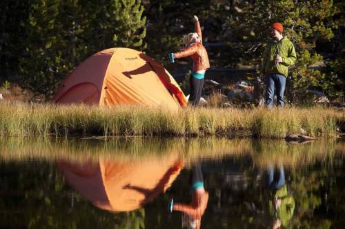 A woman stretches near an orange tent by a lake, while a man stands nearby holding a drink, surrounded by trees.