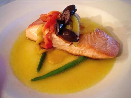 A beautifully plated salmon fillet topped with colorful peppers and olives, served in a light sauce with green beans.