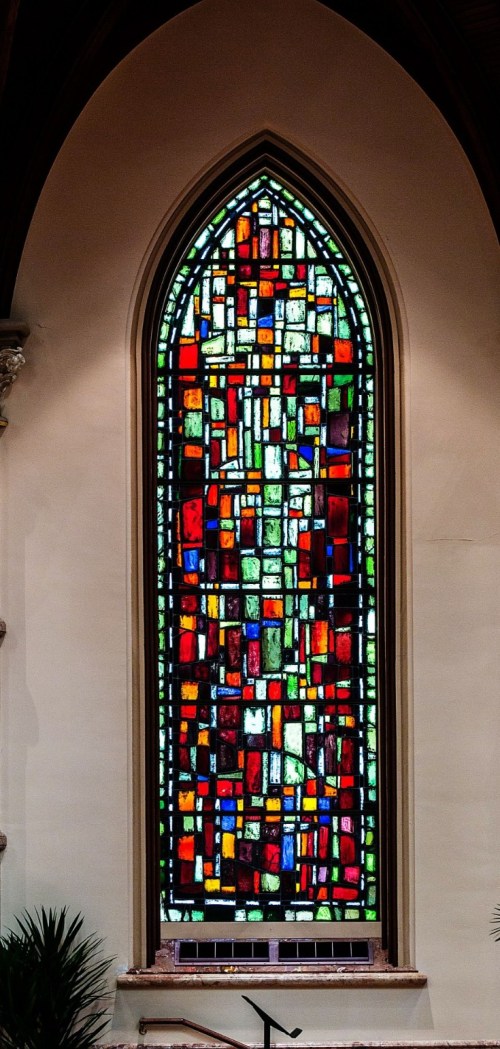 Colorful stained glass window with geometric patterns in an arched frame, set against a light-colored wall.