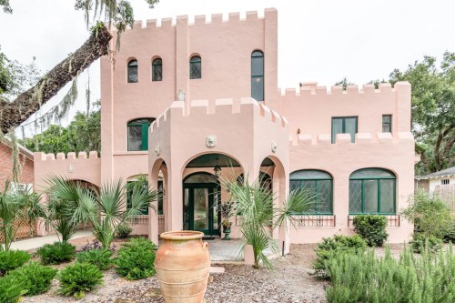 A pink castle-like house with arched windows, surrounded by greenery and a large clay pot in the front yard.