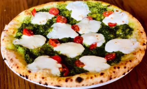 A freshly baked pizza topped with pesto, mozzarella, and cherry tomatoes on a wooden table.