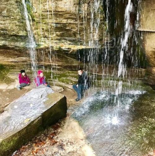 Three people sit near a waterfall in a rocky area, surrounded by lush greenery and cascading water.
