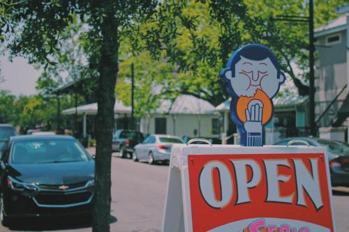 A colorful "Open" sign with a cartoon character enjoying food, set on a sunny street lined with parked cars and trees.