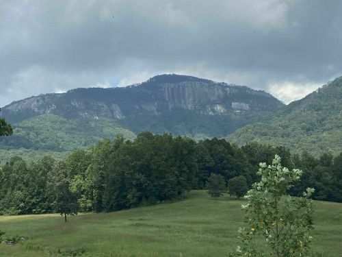 A scenic view of a mountain with a rocky face, surrounded by lush green trees and a cloudy sky.