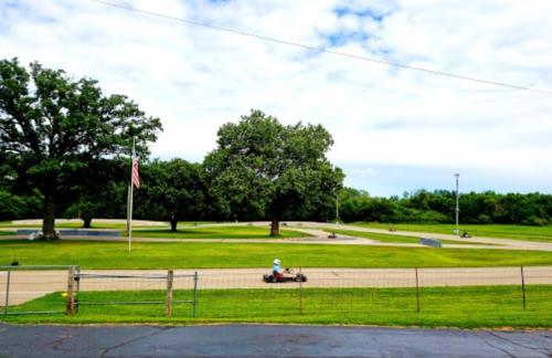 A go-kart track with several karts racing, surrounded by green grass and trees under a partly cloudy sky.