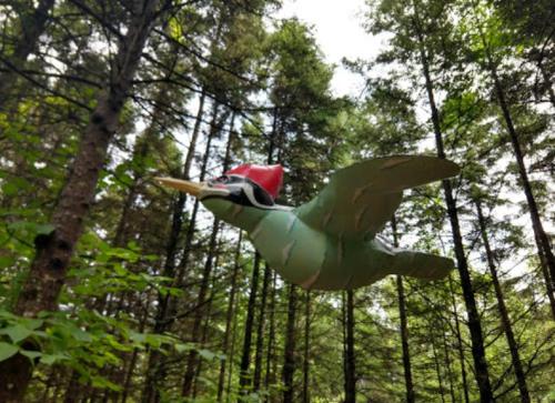 A wooden bird sculpture wearing a red hat, flying among tall trees in a forest.