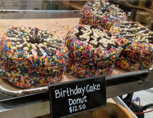 A display of colorful birthday cake donuts covered in sprinkles and topped with white chocolate pieces. Price: $12.50.