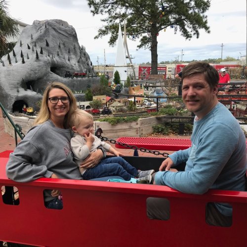 A woman holds a toddler while sitting next to a man in a red train car at a miniature train attraction.