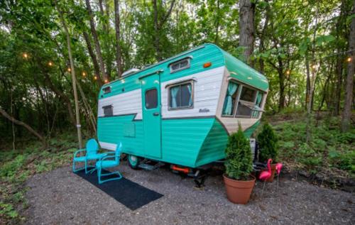 A vintage teal and white camper trailer surrounded by greenery, with outdoor seating and potted plants.