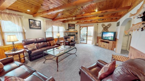 Cozy living room with wooden beams, leather sofas, a glass coffee table, and a TV, leading to a deck through sliding doors.