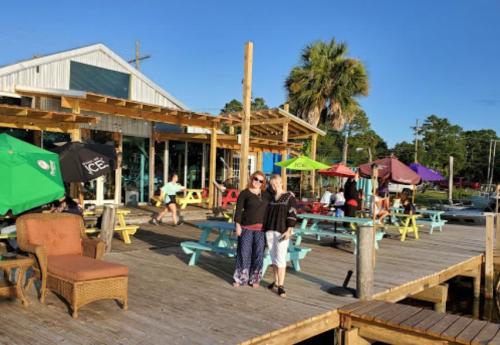 Two women pose on a wooden deck outside a lively waterfront restaurant with colorful umbrellas and seating.
