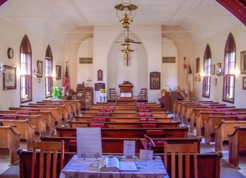 Interior of a church with wooden pews, a pulpit at the front, and decorative elements like plants and framed pictures.