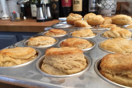 Freshly baked biscuits rising in a muffin tin, with a kitchen background featuring bottles and appliances.
