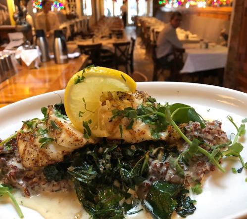 A plate of grilled fish topped with lemon and herbs, served on a bed of greens and grains, with a restaurant backdrop.