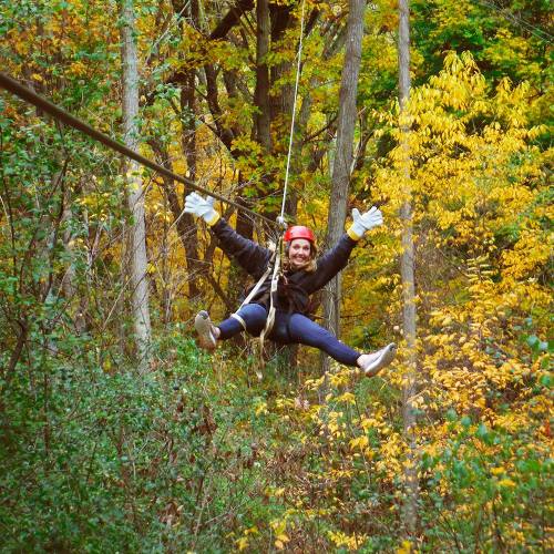 A person in a helmet and gloves zip-lining through a forest with colorful autumn foliage.