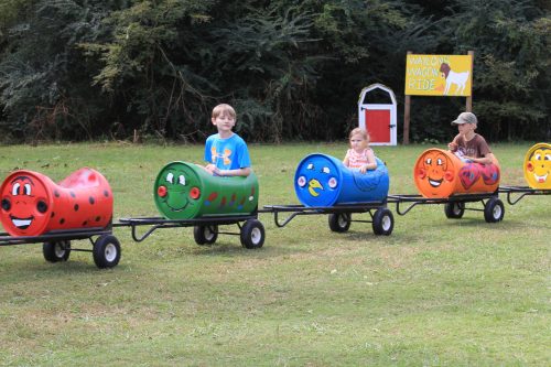 Children enjoying a colorful caterpillar ride on a grassy field, with a sign for "Waylon's Wagon Ride" in the background.