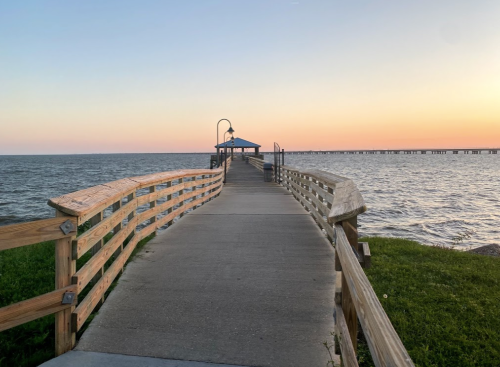 A wooden pier extends into the water at sunset, with a gazebo at the end and a calm sea under a colorful sky.