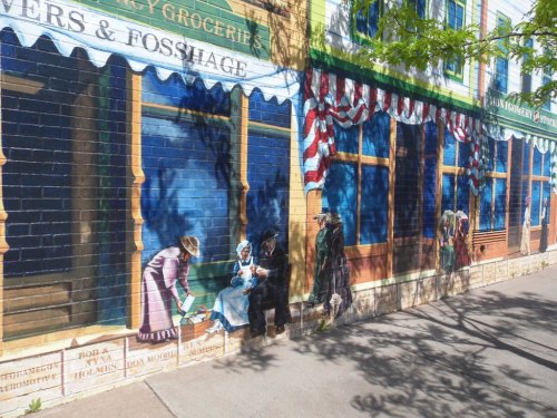 A colorful mural depicting people in vintage clothing outside a grocery store, with trees casting shadows on the wall.