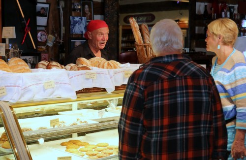 A baker in a red bandana talks to two customers in a cozy bakery filled with various breads and pastries.