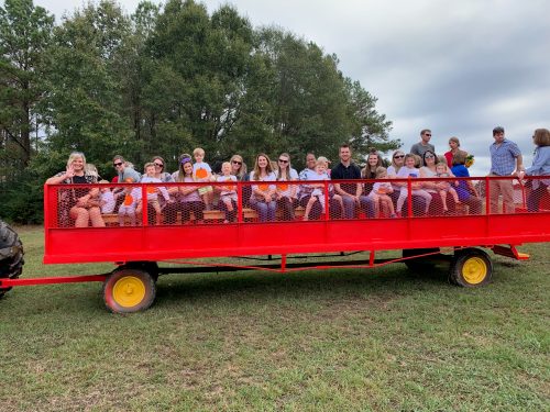 A large group of people sitting on a red wagon in a grassy field, surrounded by trees on a cloudy day.