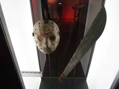 A display featuring a hockey mask and a machete, set against a lit background in a museum exhibit.