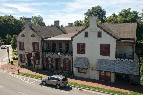 Historic building with a mix of stone and brick, featuring large windows and a shingled roof, set along a tree-lined street.