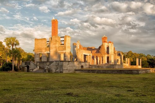 Ruins of a historic building with crumbling walls and a chimney, set against a cloudy sky and grassy landscape.