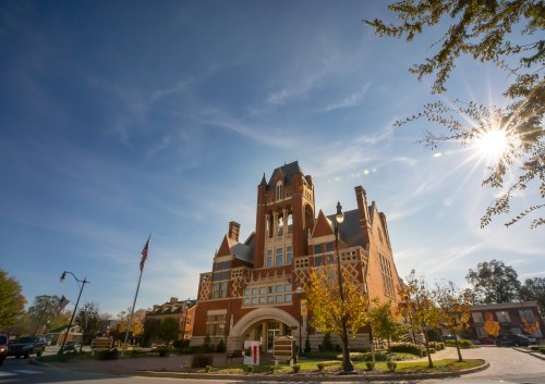 Historic brick building with a tower, surrounded by trees and a clear blue sky, sunlight shining in the background.