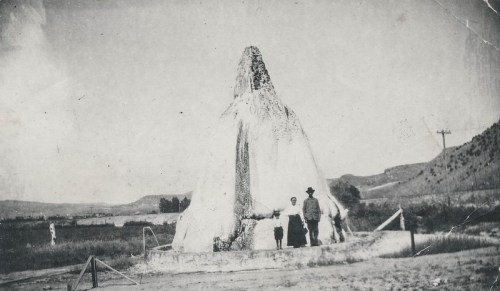 A historic black-and-white photo of a large geyser with two people and a dog standing nearby in a rural landscape.