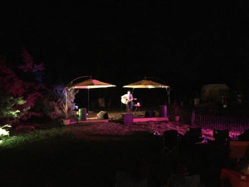 A musician performs on a dimly lit outdoor stage with colorful lights, surrounded by chairs and trees at night.