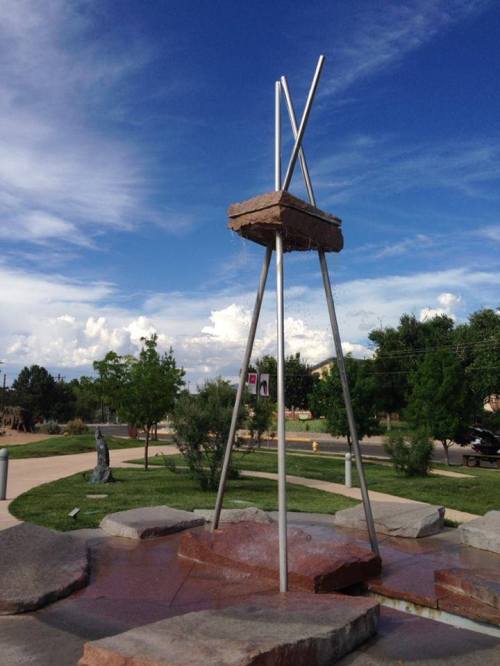 A modern sculpture with metal poles and a stone base, set against a blue sky with clouds in a park.