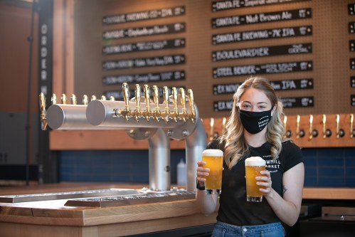 A masked bartender holds two glasses of beer in a brewery with a tap wall in the background.