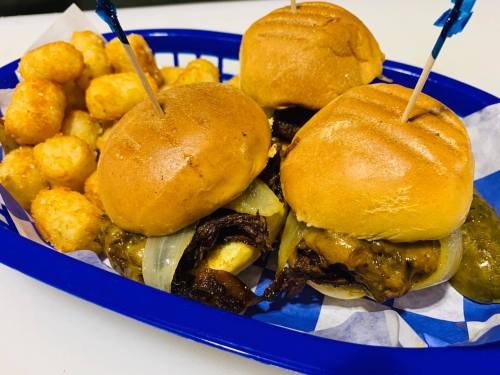 A blue basket holds two sliders with beef and onions, alongside a serving of crispy tater tots.