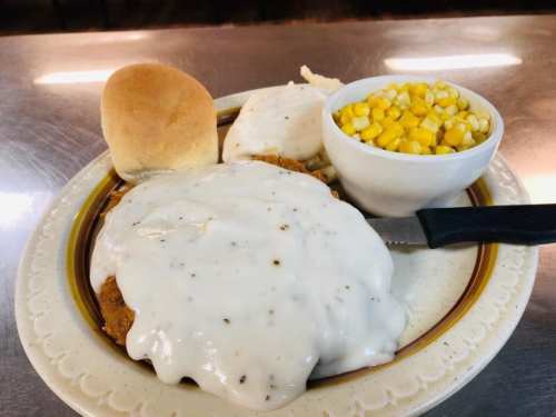 A plate of chicken smothered in white gravy, served with a roll, mashed potatoes, and a side of corn.